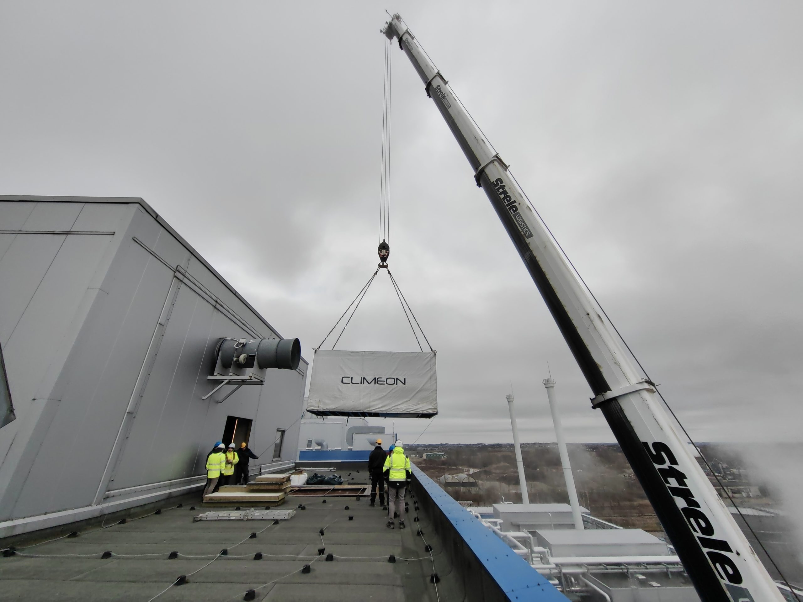 A large white crane lifts a Climeon HeatPower 300 unit above the roof of a factory as a group of workers look on.